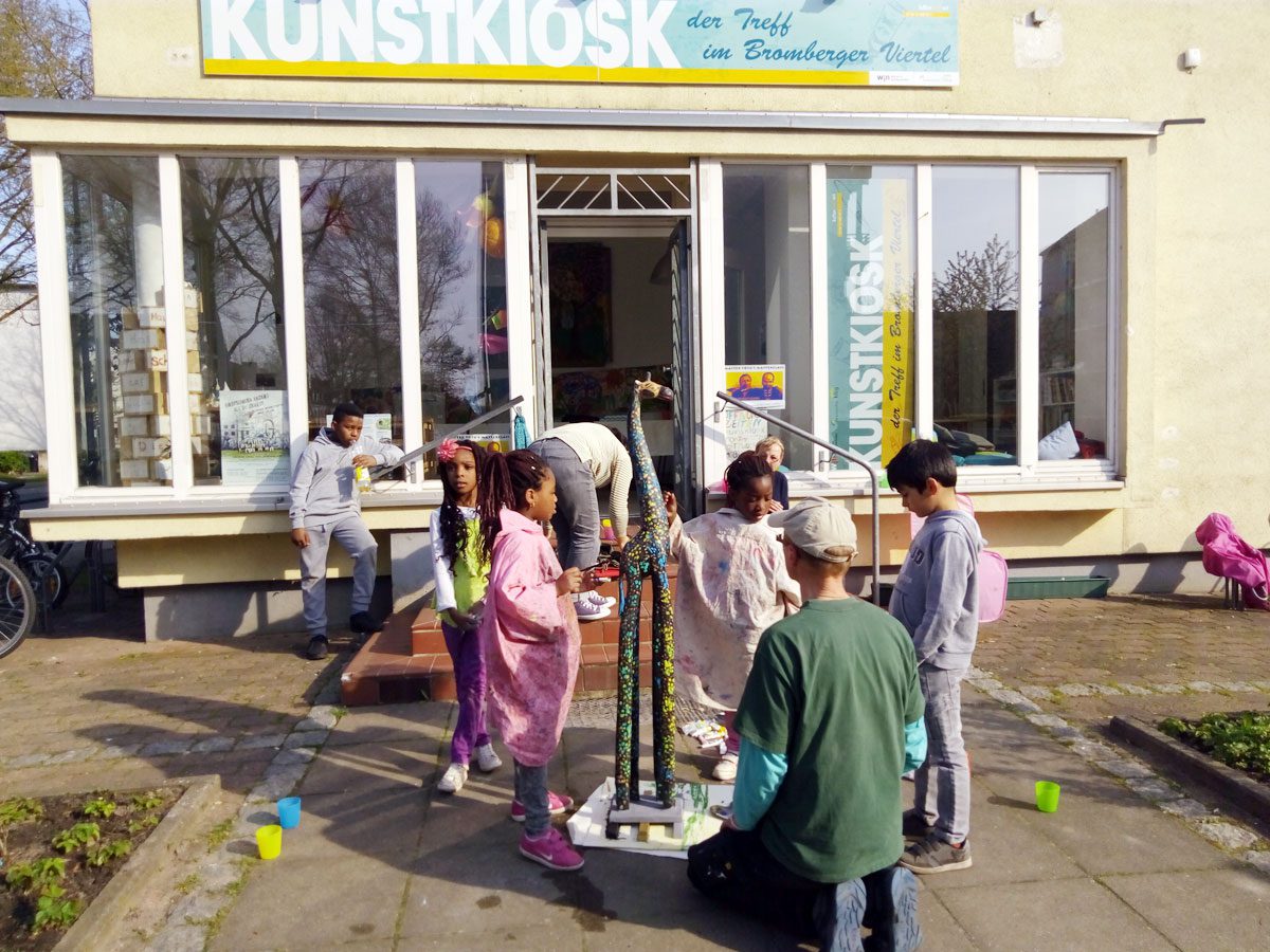 Kinder in Malkitteln spielen vor dem Kunstkiosk in der Brombergerstraße. In der Mitte steht eine Giraffen- Skulptur, die von den Kindern angemalt wird.