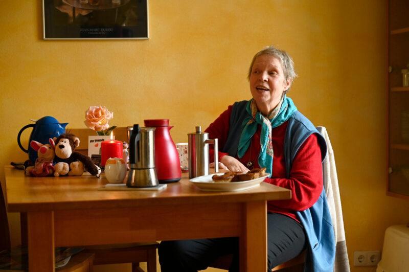 Hier seht ihr eine ältere Frau an einem gedeckten Tisch. sie erzählt gerade etwas.