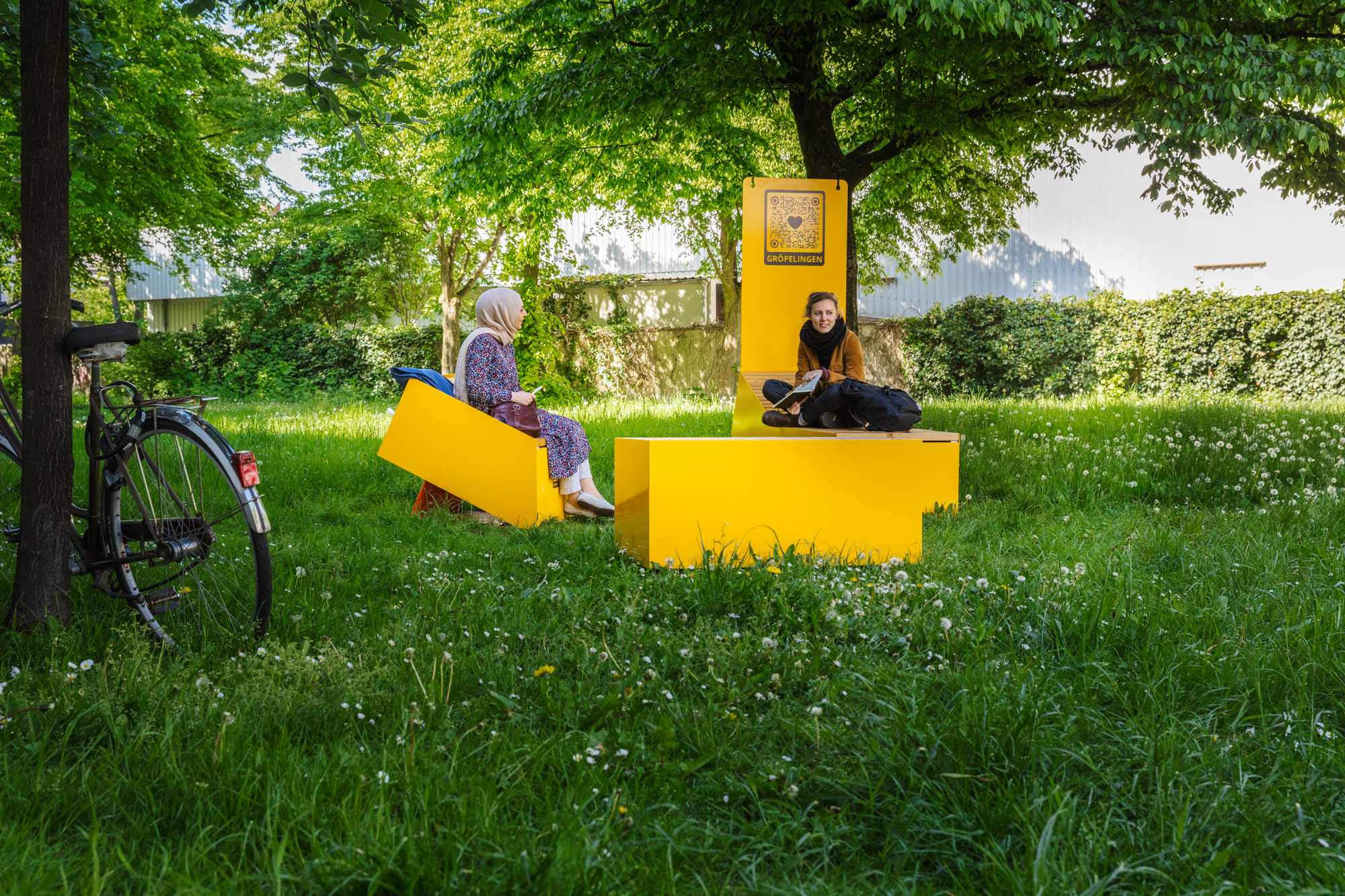 Zwei Frauen sitzen auf Stadtmöbeln im Grünzug West. Am Baum lehnt ein Fahrrad