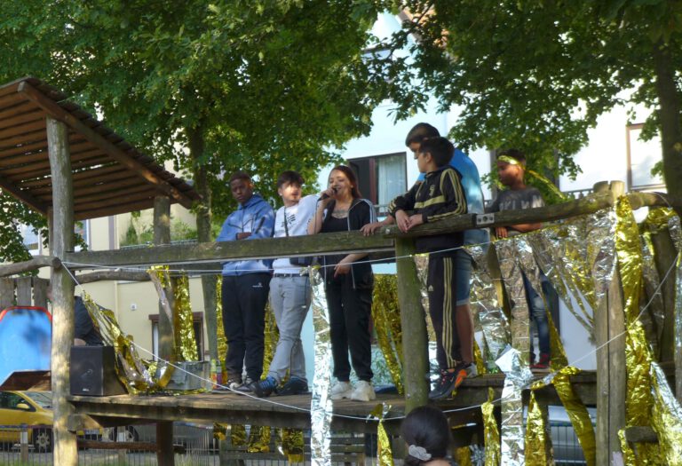 Auf einer Brücke eines Klettergerüst stehen nebeneinander fünf Jugendliche. Sie nutzen die Brücke als Bühne. Eine in der Mitte rappt in ein Mikrofon und schaut dabei nach unten ins Publikum.