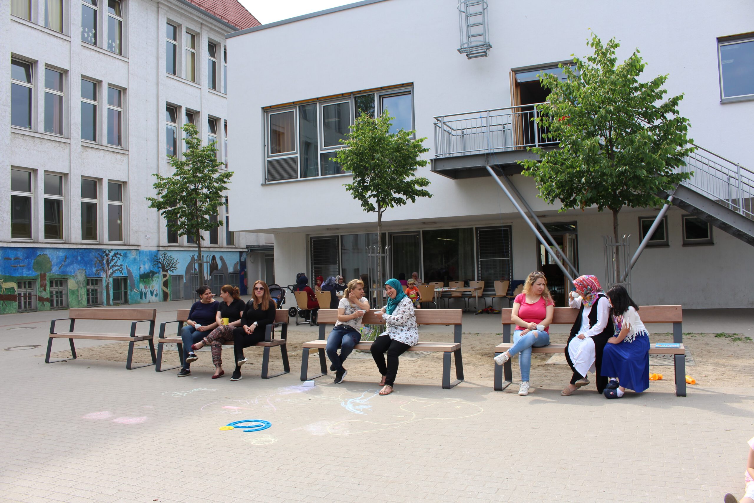 Auf dem Foto sitzen Kinde rund Erwachsene auf Bänken in einem Schulhof.