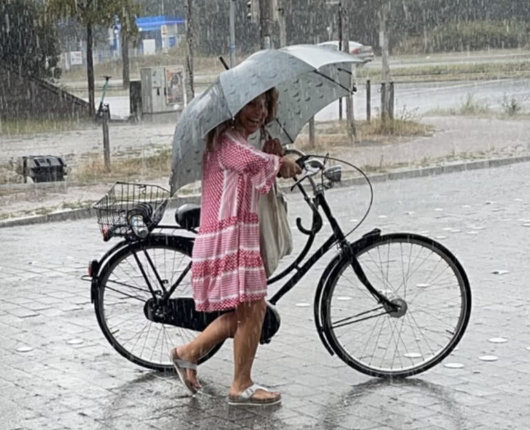Eine Frau schiebt im Regen lachend ihr Fahrrad. Sie hält einen Regenschirm in der einen Hand, in der Anderen den Fahrradlenker.