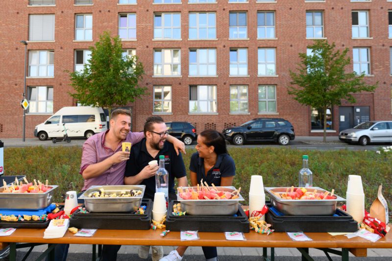 Drei Personen sitzen an lachend an einem Picknick Tisch. Das Team Rewe mit bunten Gemüsespiessen. 